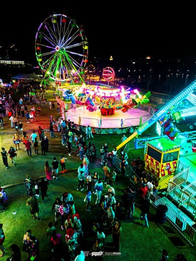 Aerial view of the lit Ferris wheel, spinning rides and crowds on the midway at night beside Armature Works, Pied Piper's Haunted Carnival 2023