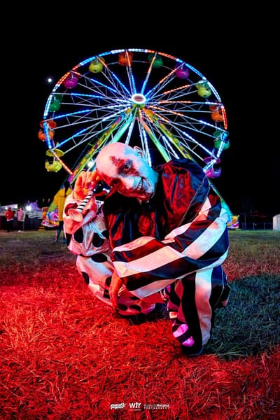 A clown in a striped costume crouching on red-lit grass in front of the glowing Ferris wheel at night, Pied Piper's Haunted Carnival 2023